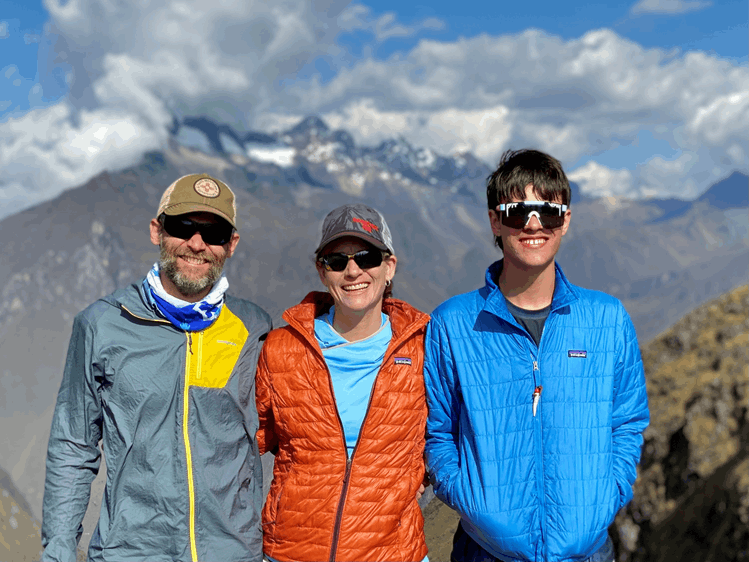 A Family of 3 wearing outdoorsy clothes with a mountain backdrop